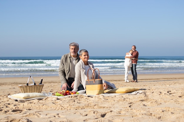 Two senior couples enjoying picnic at seashore on sunny day. Bearded man and short-haired lady sitting on blanket while another couple standing and hugging in background. Friendship, leisure concept