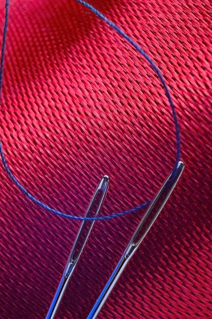 Two sewing needles with blue thread on a background of red\
fabric. close-up.