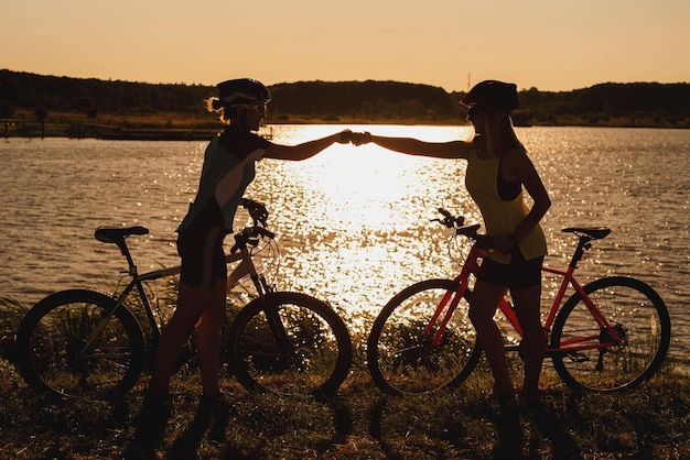 Two silhouettes of women with bike standing on lakeside and talking each other at the sunset