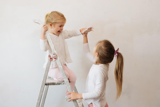 two sister child girls paint a white wall with a roller and paint at home make repairs