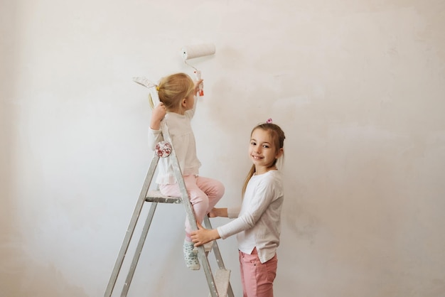 두 자매 소녀는 롤러로 흰 벽을 칠하고 집에서 페인트를 칠한다