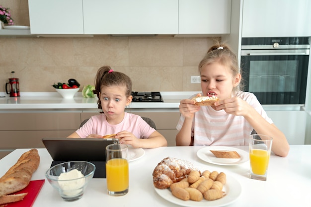 Foto due sorelle che mangiano prima colazione e che guardano insieme i cartoni animati sulla compressa, concetto nucleo familiare felice