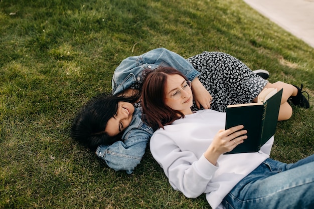 Two sisters lying on green grass in a park and reading a book