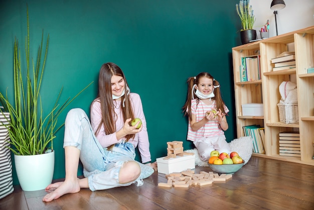 Two sisters in medical mask eating apple at home during covid quarantine.