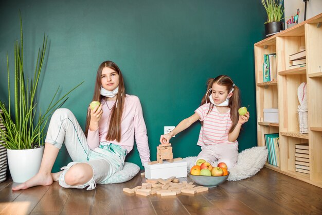 Two sisters in medical mask eating apple at home during covid quarantine.