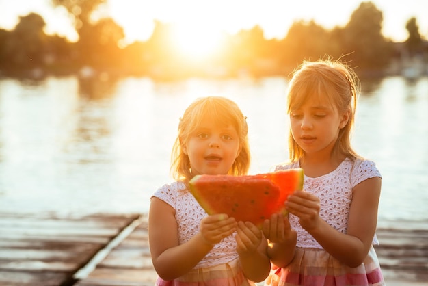 Two sisters sharing on slice of watermelon by the river at sunset