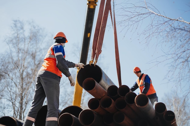 Two slingers in construction helmets and vests on street on summer day unloaded metal pipes Real scene