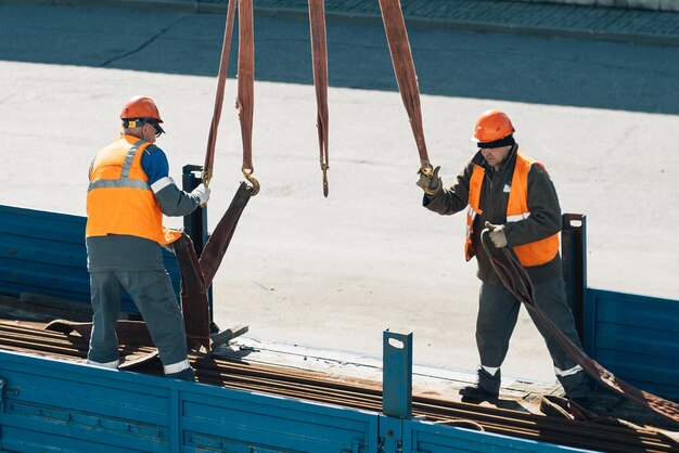 Two slingers hook cargo to slings in back of truck Real workers unload cargo Slingers in construction helmets and vests at work on sunny day Production background