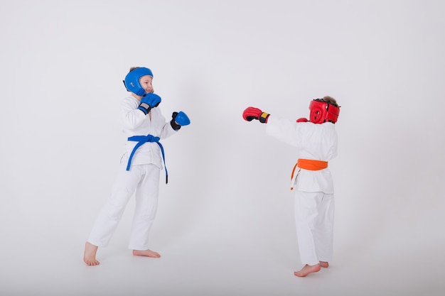 Two small boy in a white kimono and wearing a helmet with gloves fighting on white wall with a copy of the space