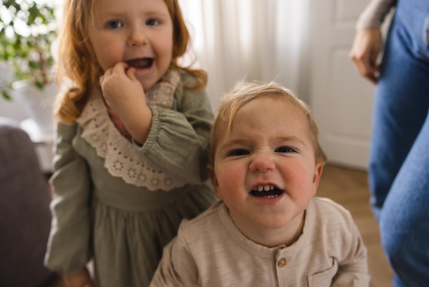 Two small fairskinned children blond boy and redhaired girl grimace at camera in room Kinship concept