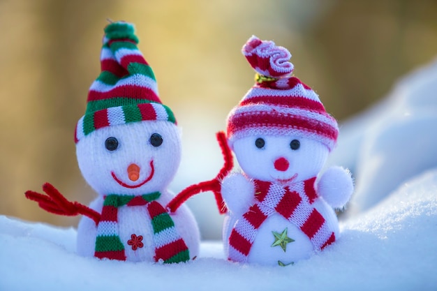 Two small funny toys baby snowman in knitted hats and scarves in deep snow outdoors on bright blue and white copy space background. Happy New Year and Merry Christmas greeting card.