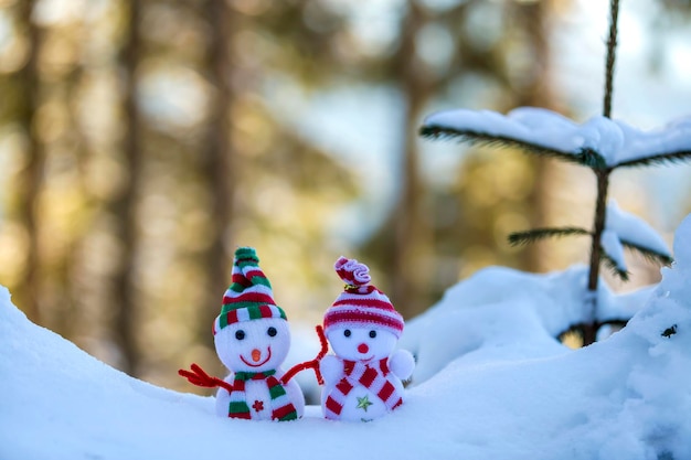 Two small funny toys baby snowman in knitted hats and scarves in deep snow outdoors near pine tree branch Happy New Year and Merry Christmas greeting card