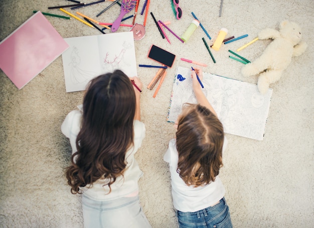 Due bambine sdraiato sul tappeto e disegno. hanno matite e carta bianca su tutto il pavimento.