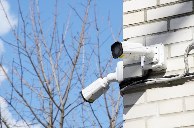 Две маленькие белые камеры видеонаблюдения на углу фасада многоэтажного кирпичного дома.