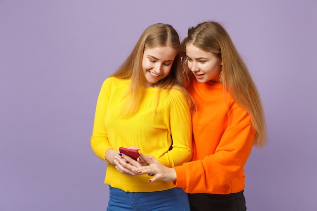 Two smiling amazed young blonde twins sisters girls in vivid clothes using mobile phone typing sms message isolated on violet blue wall . People family lifestyle concept. 