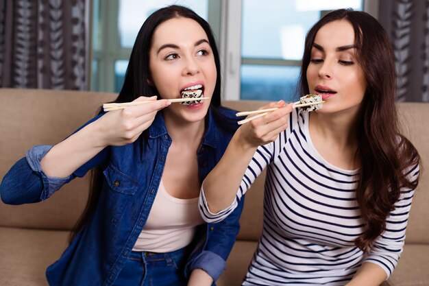 Two smiling beautiful girl friends in modern casual clothes sitting on the couch eating vegan rolls and communicate