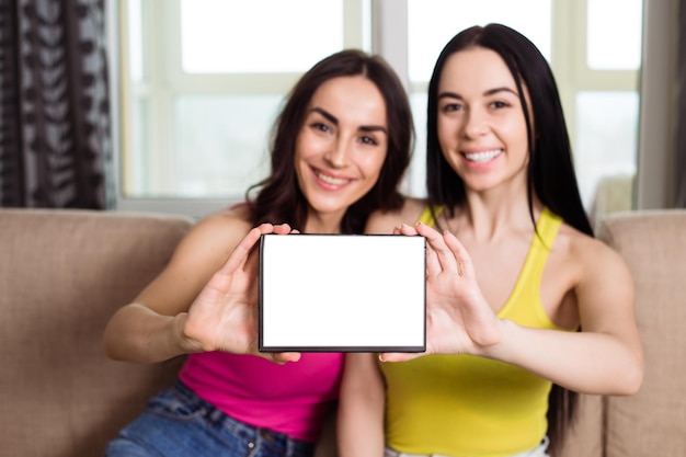 Two smiling beautiful young girlfriends in casual clothes hold a white sheet for advertising sitting on the sofa at home