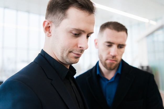 Two smiling businessmen in airport, discussing important project and looking at phone screen.