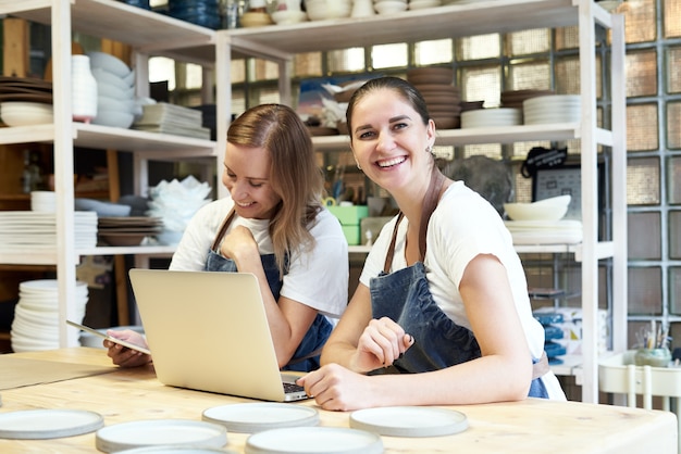 Две улыбающиеся женщины-предпринимательница с ноутбуком в мастерской ремесленника.