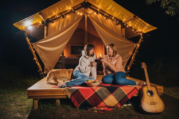 가을 저녁 모닥불에 아늑한 글램핑 텐트에 앉아 와인을 마시고 과일을 먹고 있는 웃고 있는 두 여자 친구 야외 휴가 및 휴가 라이프스타일 컨셉을 위한 럭셔리 캠핑 텐트