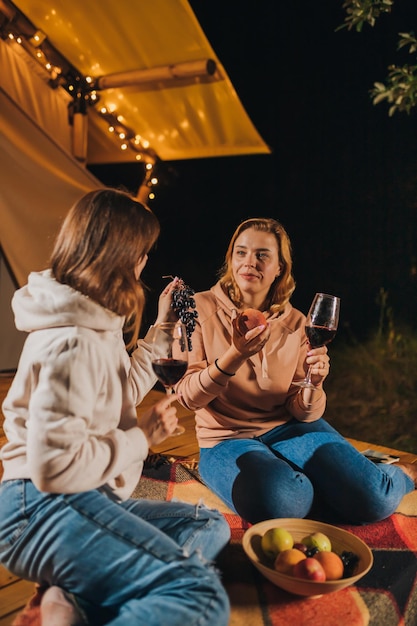 Two smiling female friends drinking wine and eating fruits\
sitting in cozy glamping tent in autumn evening bonfire luxury\
camping tent for outdoor holiday and vacation lifestyle\
concept