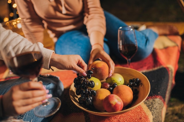 Two Smiling female friends drinking wine and eating fruits sitting in cozy glamping tent in autumn evening bonfire Luxury camping tent for outdoor holiday and vacation Lifestyle concept