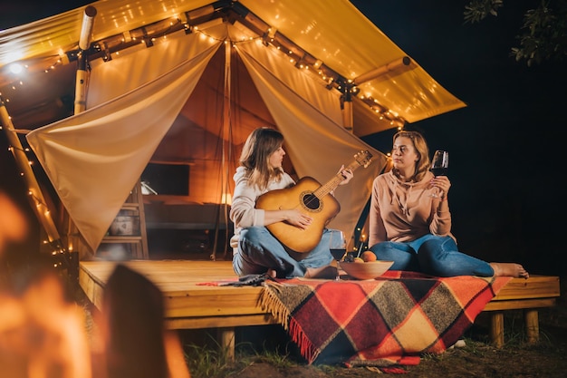 Two smiling female friends drinking wine and play guitar sitting in cozy glamping tent in autumn evening bonfire luxury camping tent for outdoor holiday and vacation lifestyle concept