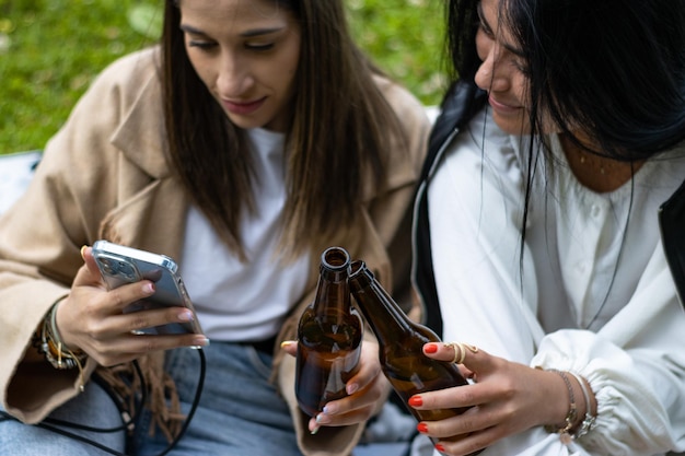 Two smiling female friends toasting with beer sitting on the grass Lifestyle of young female friends sitting on the grass toasting with beer bottles