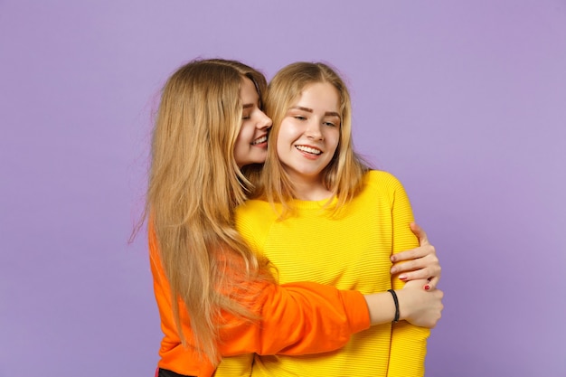 Two smiling funny young blonde twins sisters girls in vivid colorful clothes hugging, looking aside isolated on pastel violet blue wall  . People family lifestyle concept. 