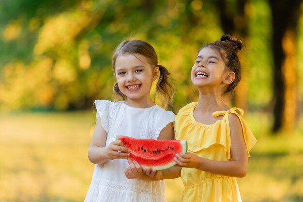 Two smiling girls are holding one slice of watermelon in their hands Kids eat fruit outdoor