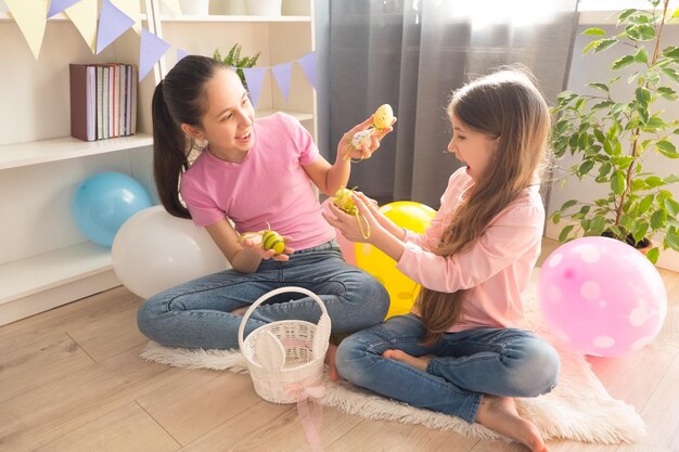 Two smiling girls collect eggs in basket celebrating Easter holiday room decorated with balloons