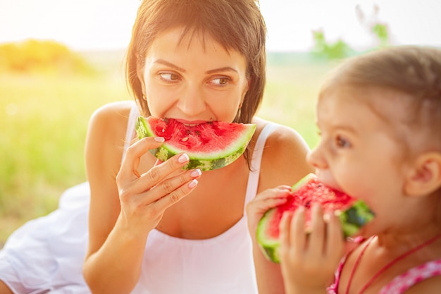 Two smiling girls eats slice of watermelon outdoors on meadow. Mother and daughter spend time together. Diet, vitamins, healthy food concept.