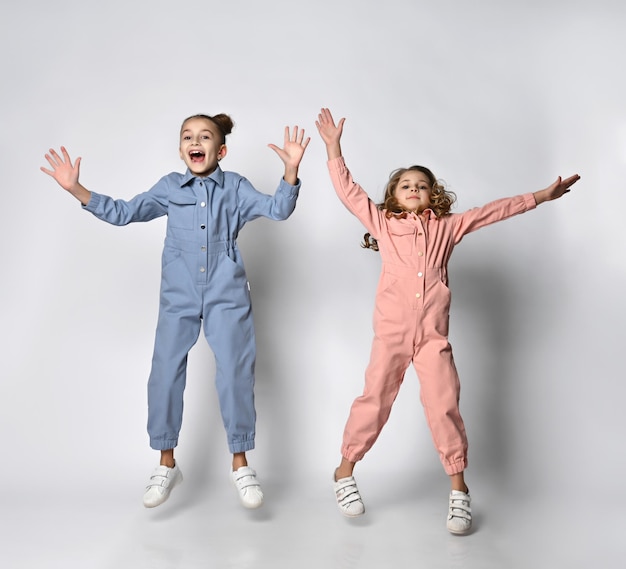 파란색과 분홍색 면 작업복을 입은 웃고 있는 두 여자 친구 자매와 주머니가 즐겁게 점프하고 뿌리다