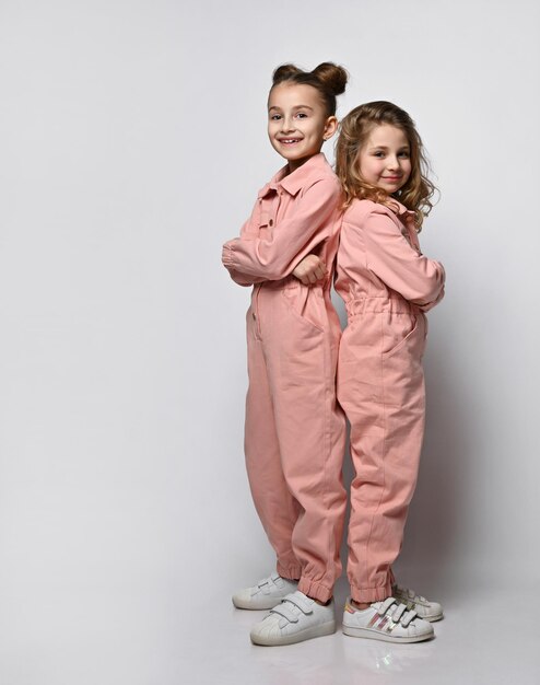 Two smiling girls friends sisters in pink cotton overalls with pockets stand close to each other