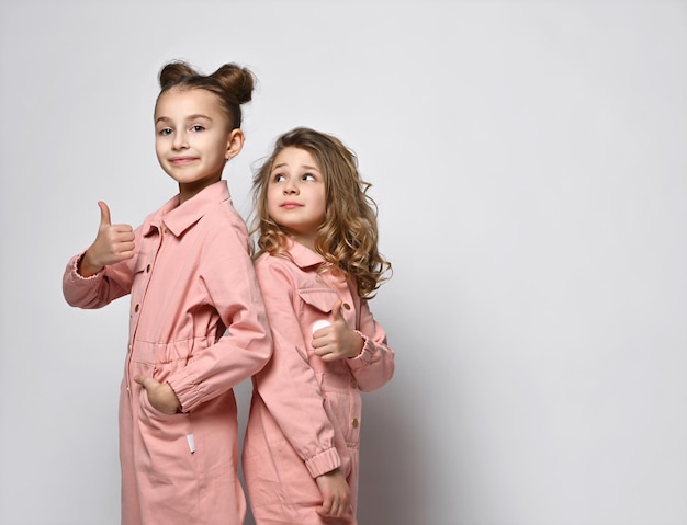 Two smiling girls girlfriends sisters in pink cotton overalls with pockets stand close to each other