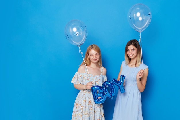 Two smiling girls in light dress holding blue balloons and balloon with the inscription boy isolated blue background