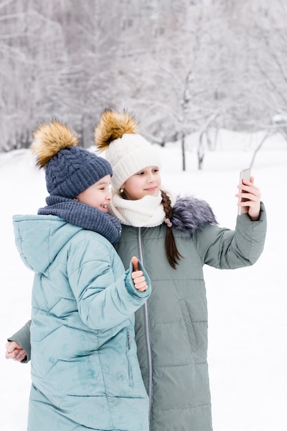 暖かいコートと帽子をかぶった2人の笑顔の女の子が、雪に覆われた冬の公園でスマートフォンで自分撮りをしますライフスタイルテクノロジーの使用垂直方向のビュー