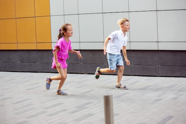 Two smiling kids, boy and girl running together in town, city in sunny day. Concept of childhood, happiness, sincere emotions, carefree lifestyle. Little caucasian models in bright clothes.