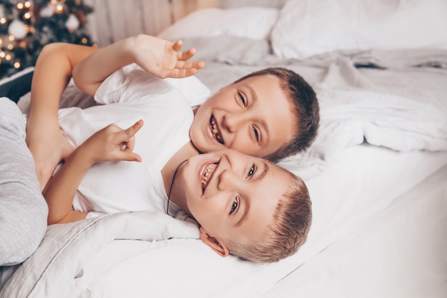 Two smiling kids having fun, laughing and hugging on the white bed with Christmas tree