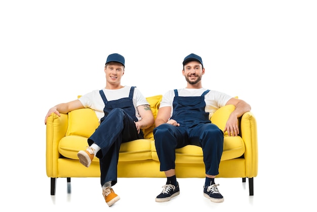 Two smiling movers in uniform sitting on yellow sofa and looking at camera isolated on white