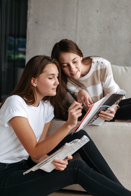 Two smiling teenage girls reading magazines while sitting on a couch at home