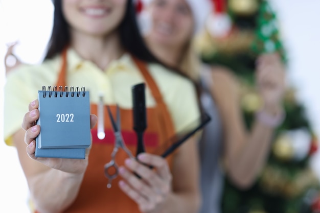 Two smiling women hairdressers in uniforms hold 2022 calendar and scissors with a comb. Barber profession training concept