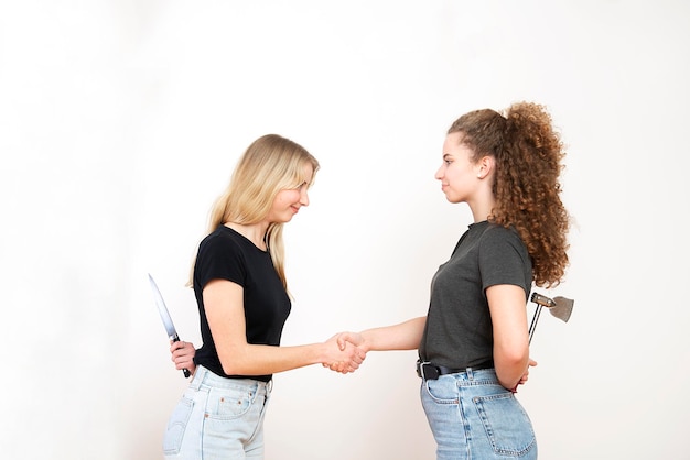 Photo two smiling women standing face to face and hiding knife and axe behind back isolated on white