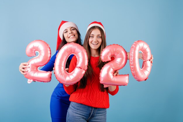 Two smiling women wearing santa hats holding 2020 balloons for new year holiday isolated over blue