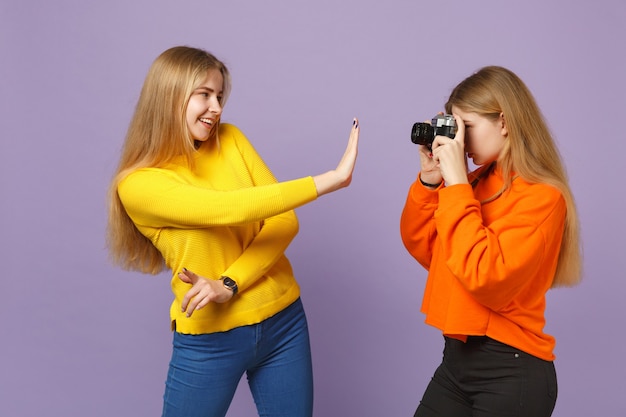 Two smiling young blonde twins sisters girls in colorful clothes taking pictures on retro vintage photo camera isolated on violet blue wall. People family lifestyle concept. 