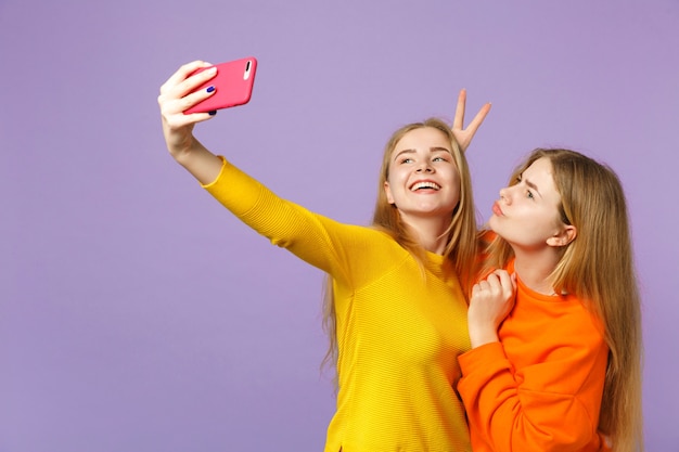 파스텔 바이올렛 블루 벽에 고립 된 휴대 전화에 셀카 촬영을 하 고 화려한 옷에 두 웃는 젊은 금발 쌍둥이 자매 여자. 사람들이 가족 라이프 스타일 개념.