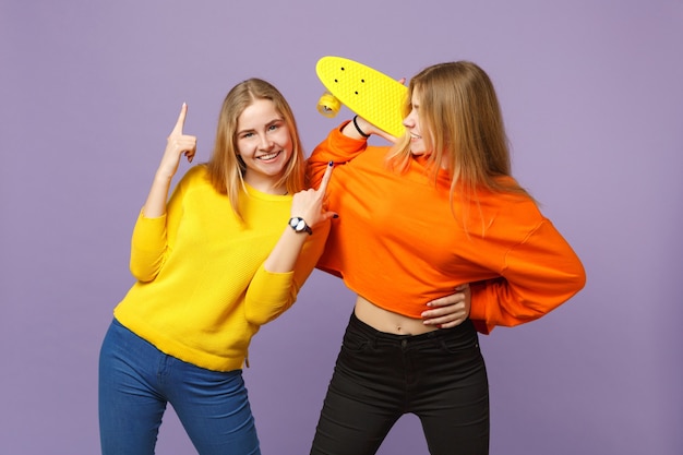 Two smiling young blonde twins sisters girls in vivid clothes pointing index fingers up, hold yellow skateboard isolated on violet blue wall. People family lifestyle concept. 