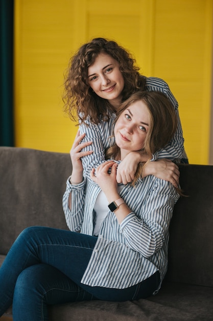 Две улыбающиеся молодые девушки в полосатых рубашках, джинсах и кроссовках позируют на диване. студийная съемка.