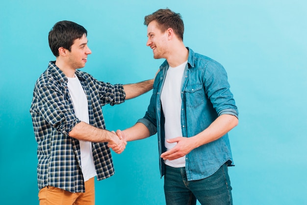 Two smiling young men shaking hands against blue background