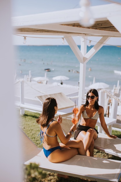 Two smiling young women in bikini enjoying vacation on the beach while drinking cocktail